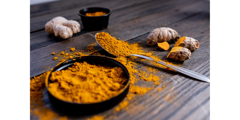 A styled photo of turmeric powder in a wooden bowl with a silver spoon, accompanied by whole, fresh turmeric roots, highlighting the vibrant golden color and earthy texture. For Noshejan's blog post titled A Love Letter to the Golden Spice.